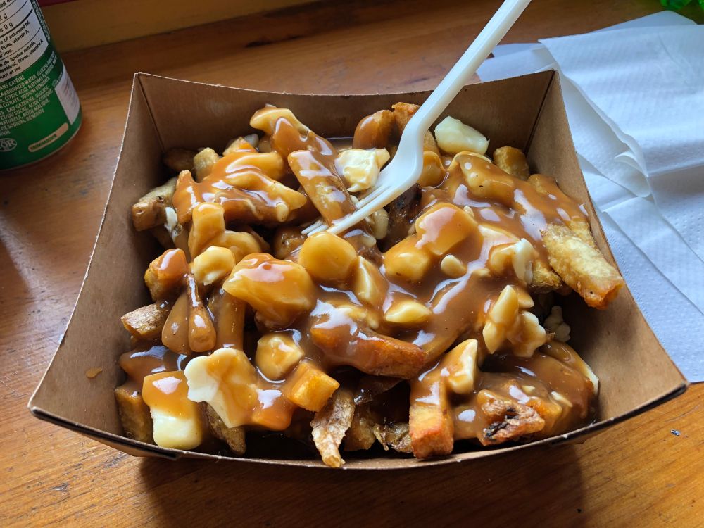A photograph of poutine in a disposable bowl on a wooden table in a restaurant.
