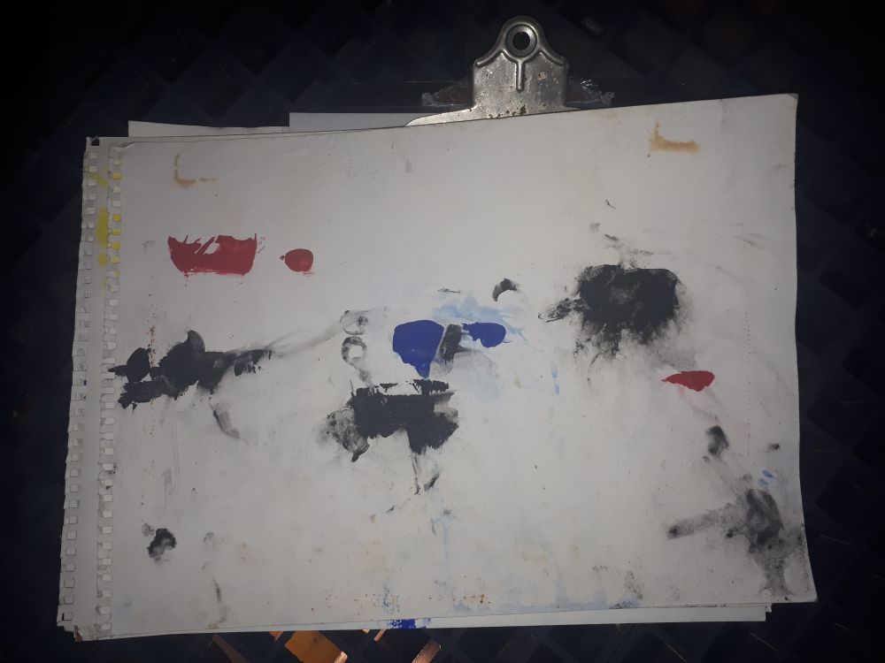 A piece of paper with splattered paint over it attached to a clipboard, it is red and blue and black.