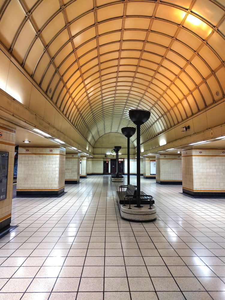 Photo of the waiting area of an old tube station. The platforms for both directions are visible from the middle. There are seats with lights pointing up to the ceiling.