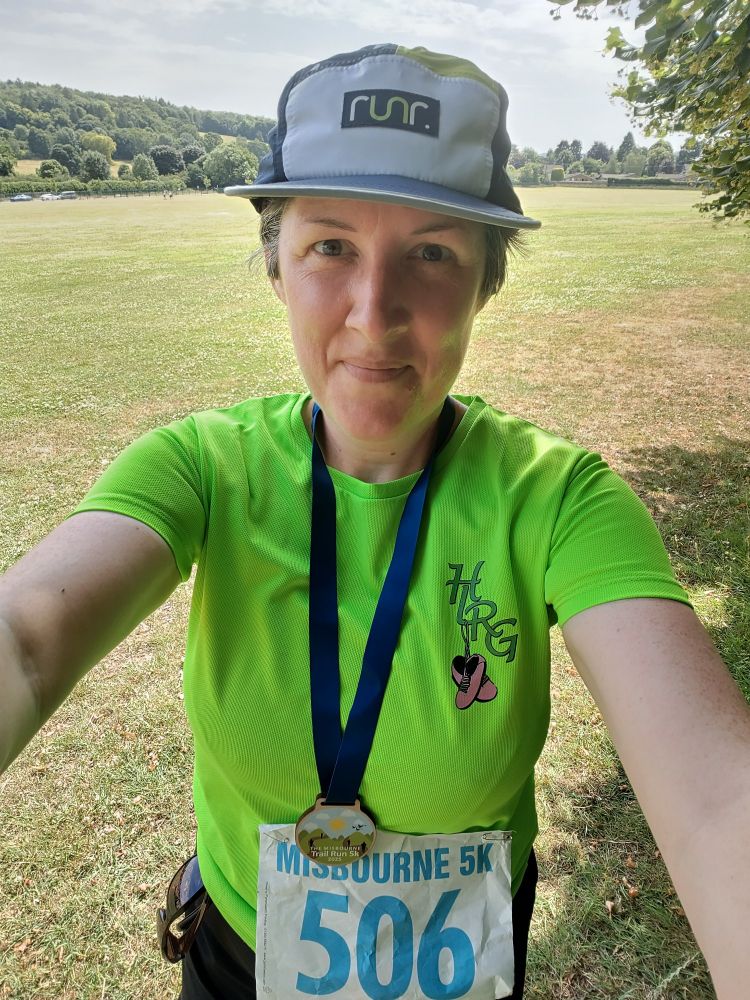 Selfie of a very satisfied, but hot, woman who has just confirmed l finished a hilly 5k trail race. She is wearing a peaked hat, green shirt and a finishers medal.