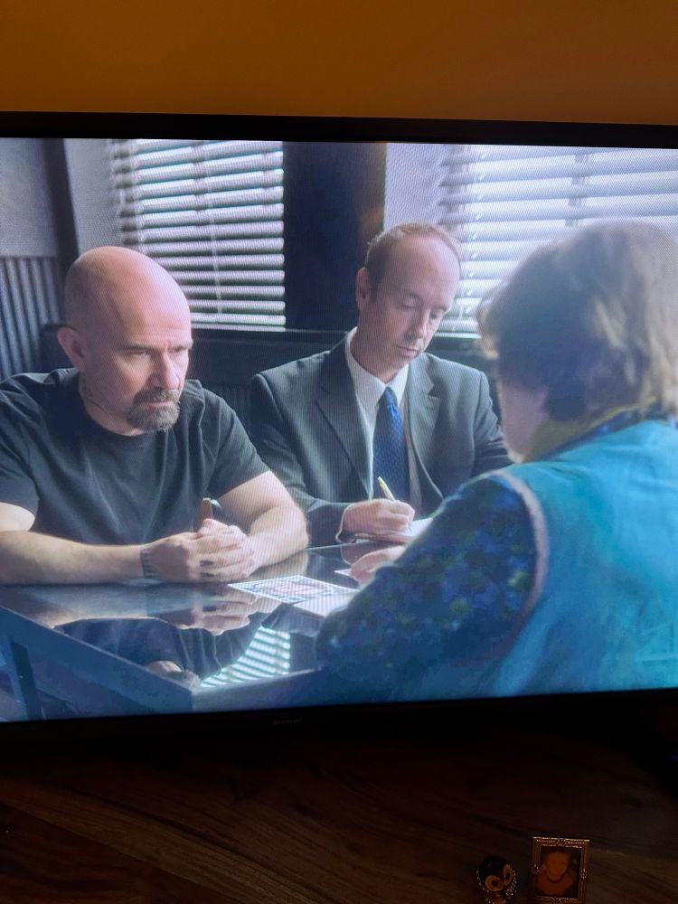 A photo taken of the programme Vera on my TV showing an interrogation scene with Vera, wearing shades of light and dark blue, with her back to the camera on the right, and my husband Chris, black tee, skinhead, dark beard and looking just like Walter White in Breaking Bad facing her, with a lawyer in a suit to the right
