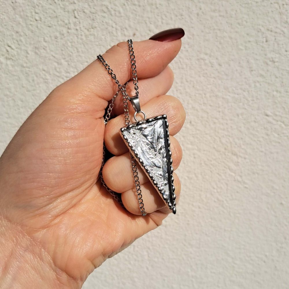 Pendentif en forme de triangle que je tiens par sa chaine.
Il est composé de verre miroir coloré argenté et encadré d’étain pur selon la méthode du vitrail tiffany.