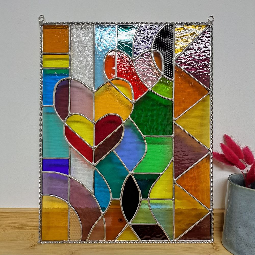 Un vitrail très coloré et abstrait, composé de formes géométriques, de ronds, de vagues et de 3 coeurs. Il est encadré de tiges torsadées. 

Il est posé sur une étagère. 