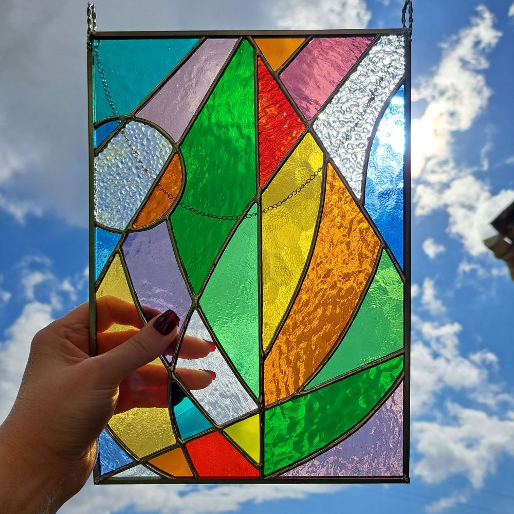 Un vitrail tiffany de forme rectangulaire. Il est composé de verres colorés, texturés, de formes géométriques. Certains y voient un bateau mais il s'agit d'une feuille stylisée. Je le tiens devant un ciel bleu nuageux.