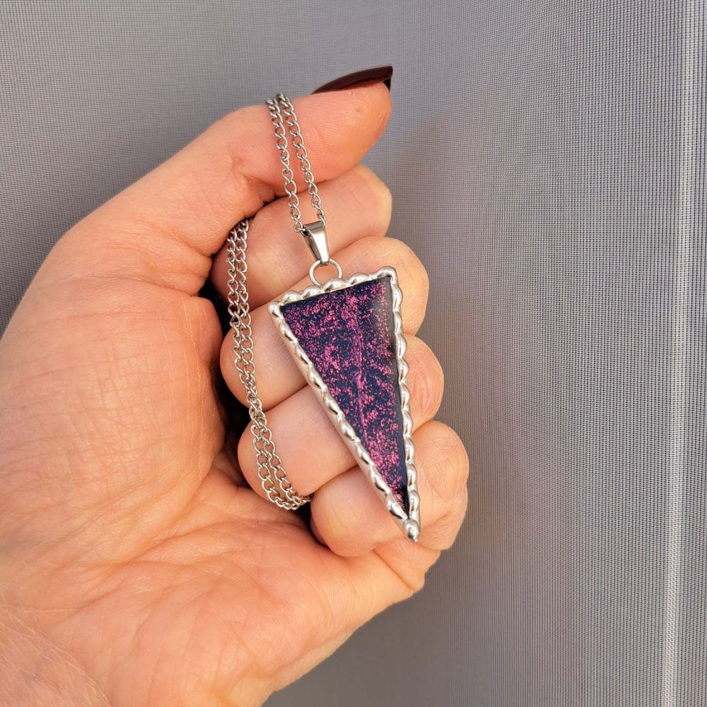 Pendentif en forme de triangle que je tiens par sa chaine.
Il est composé de verre miroir coloré (dans les tons violet/bronze) et encadré d’étain pur selon la méthode du vitrail tiffany.