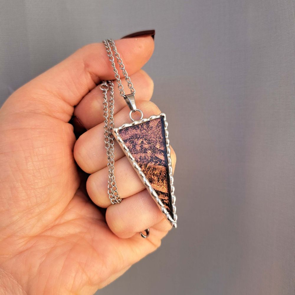 Pendentif en forme de triangle que je tiens par sa chaine.
Il est composé de verre miroir coloré (dans les tons bronze) et encadré d’étain pur selon la méthode du vitrail tiffany.
