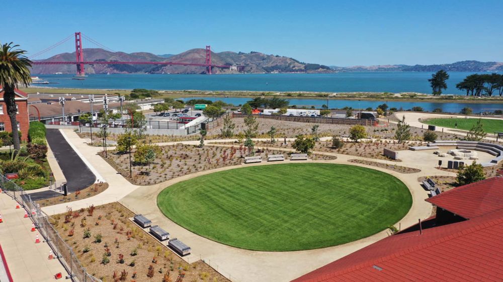 a photo of the new presidio tunnel tops park that shows the park from an elevated perspective. there is a round green grass area surrounded by benches and pathways. the golden gate bridge and the water of the bay are in the background beneath a clear blue sky.
