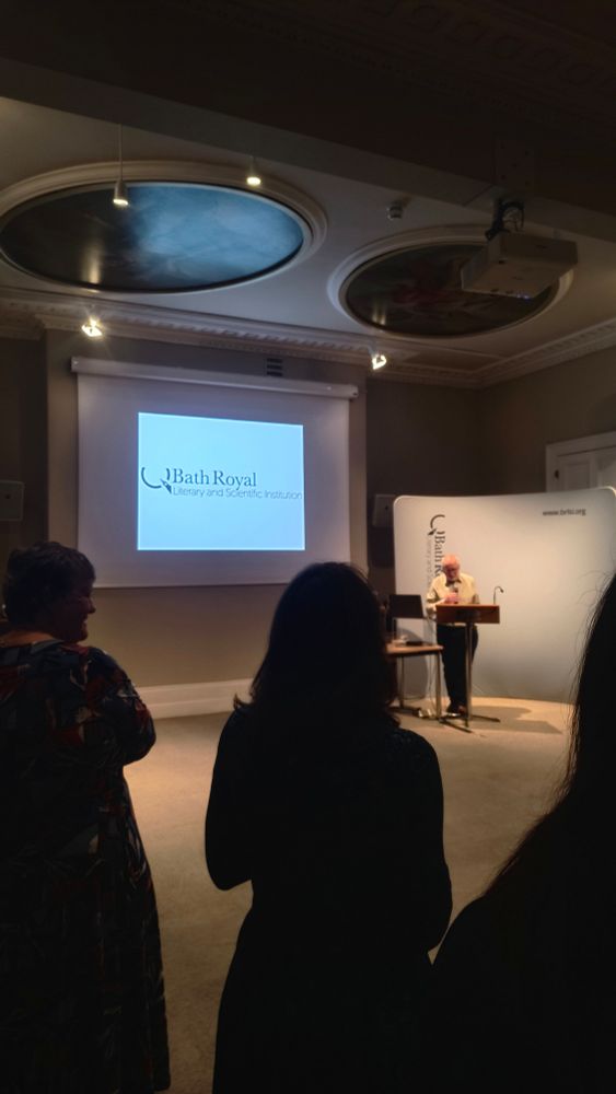 At a podium infront of a silhouetted crowd Jon Lorie, the new course director of MA Nature & Travel Writing Bath Spa, introduces speeches of success, rallies and appreciation. At Bath Royal Literary & Science Institute (written on a power point behind).