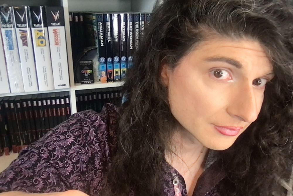 Krisis in their home studio earlier this week recording their Top 60 Comics Series of 2025 (so far) stream. They are turned towards the camera in three quarter profile. They are wearing their hair long in curls in a black shirt with a purple paisley print. Behind them, shelves of books hold releases from Valiant, DC, and Marvel.