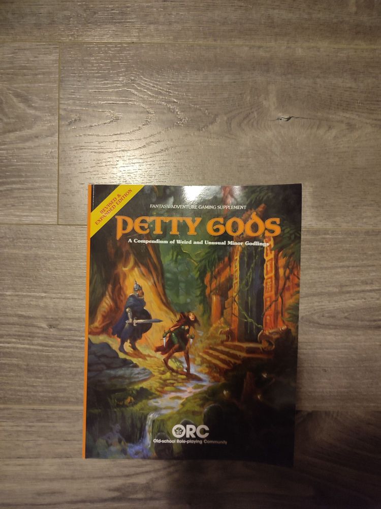 A glossy, paperback book titled Petty Gods Expanded and Revised Edition is on a grey, wooden floor with a light directly above it. A shadow of the photographer is visible. It is by the Old School Roleplaying Community. It has the art of a fighter-like figure and a rogue-like figure walking through a forest when they encounter an ancient stone ruin. This book is a fantasy adventure gaming supplement and it is a compendium of weird and unusual minor godlings. 