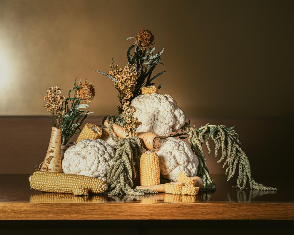 A still life photo of a artistically crafted scene made up of vegetables and flowers. The scene is set in light yellow and some green tones. The vegetable sculpture is made up of corn cobs, cauliflower and several other plants