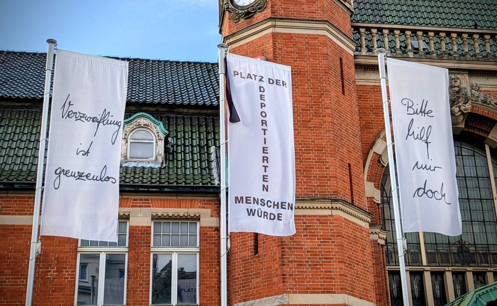 Drei weiße Fahnen an Fahnenmasten vor dem Hintergrund Bahnhofsgebäude Lübeck Hauptbahnhof.

Aufschrift auf den Fahnen: 

Verzweiflung ist grenzenlos 

Platz der Deportierten - Menschenwürde 

Bitte hilf mir doch. 

