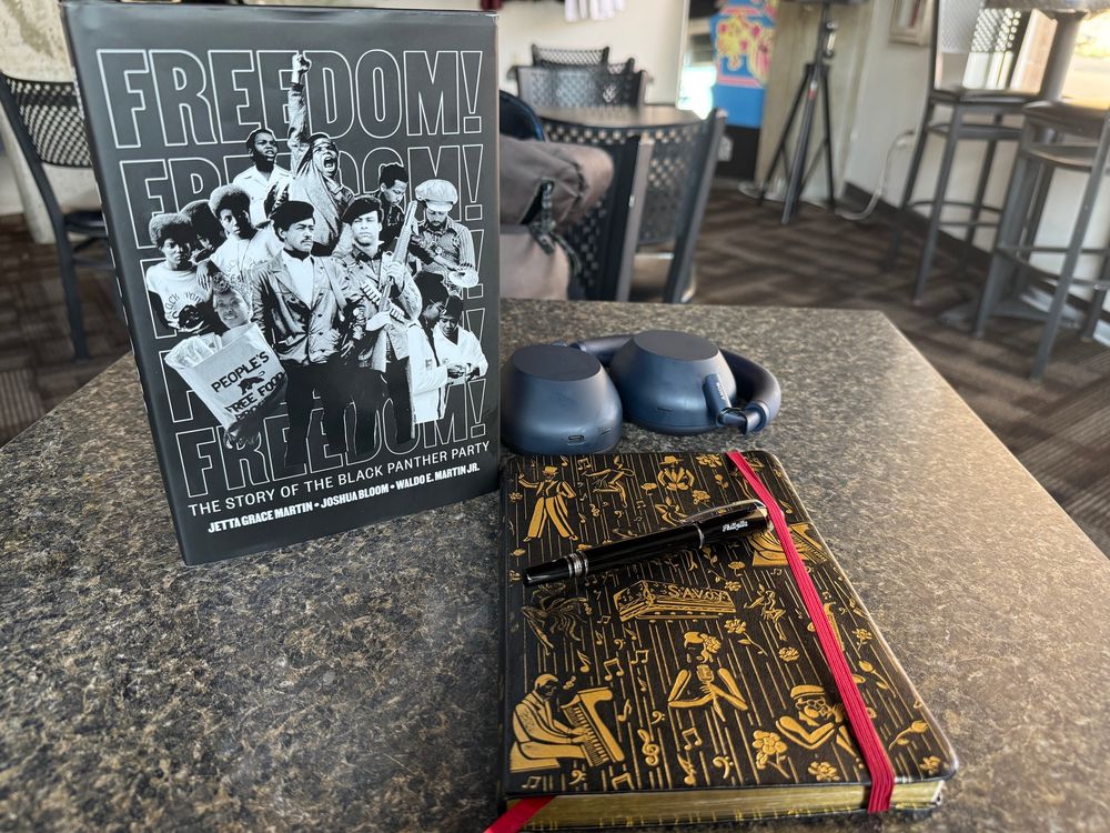 Book: Freedom: The story of the black panther party, headphones, a journal, and a pen on top of a coffee table. 