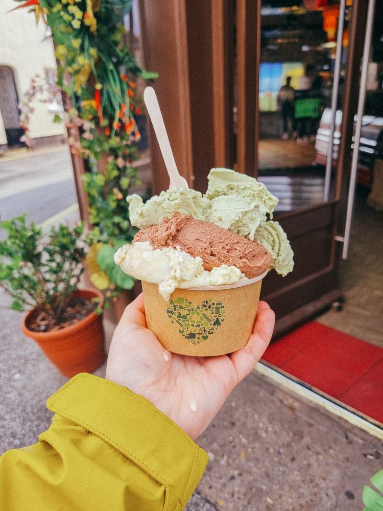 a photo of my hand holding a large tub of ice cream outside of the ice cream shop. I have 3 flavours. on the bottom is tiramisu, in the middle is chocolate and on top with a wooden spoon is pistachio 