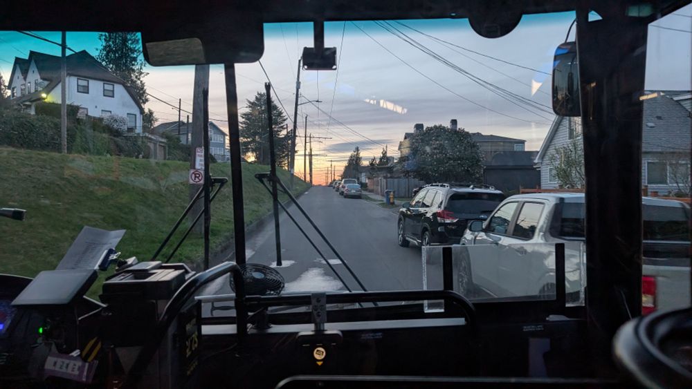 A street split in two where the southbound lane is significantly higher than the northbound lane. Picture is taken looking out the front of a bus. The sun is setting, and there is trolleybus wire strung over the street 