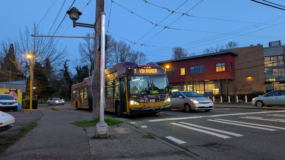 A full shot of a 60 foot trolleybus with destination sign "7 Via Rainier". It is now dark 
