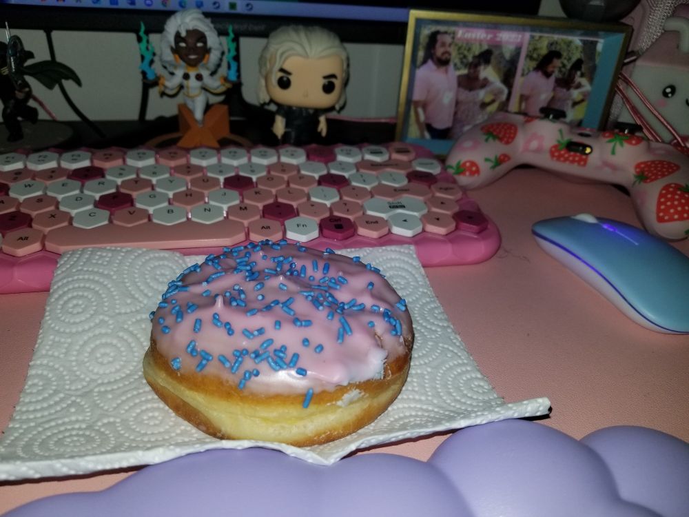 Cotton candy donut with pink icing and blue sprinkles on a paper towel on a pink gaming desk with a pink keyboard, blue mouse, and purple cloud arm rest.