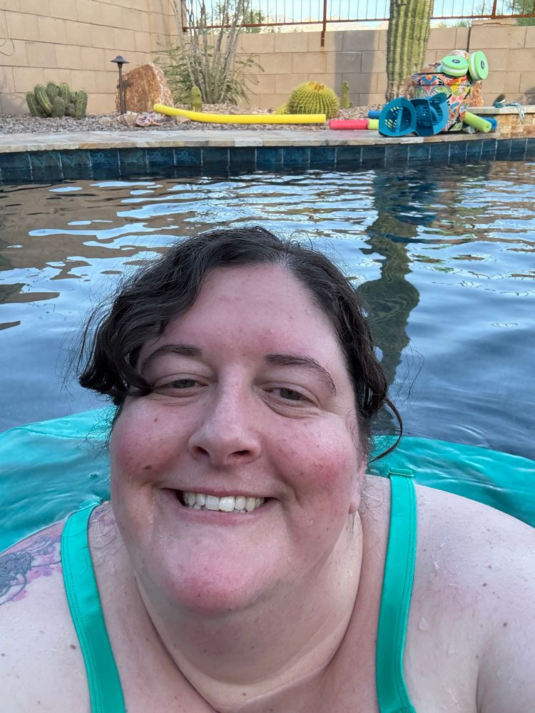 A selfie of the author in the pool. Her hair is pulled back and she’s wearing a turquoise swimsuit, which is billowing in the water behind her. There is part of a tattoo visible on her right shoulder, and a saguaro is reflected in the water behind her. 