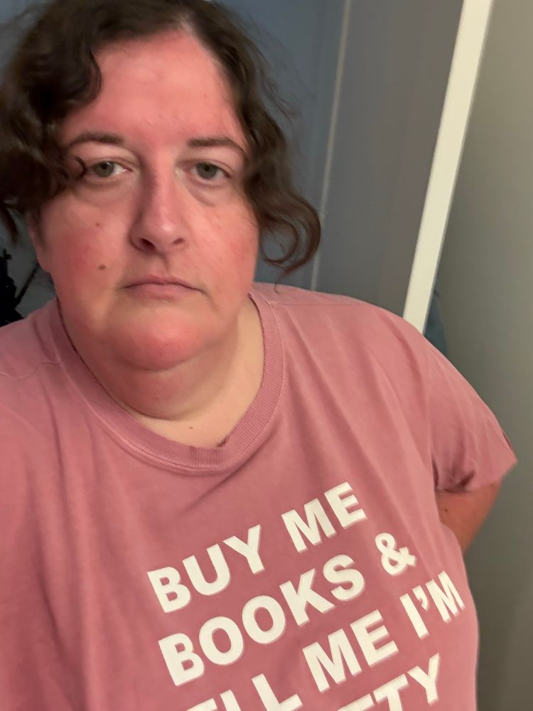 A selfie of the author in a pink tee shirt that says “buy me books and tell me I’m pretty”