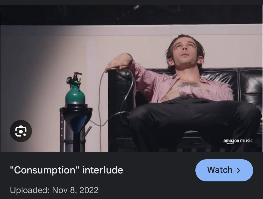 Print screen de um frame do vídeo feito da interlude do show At Their Very Best, com Matty Healy sentado em um sofá preto, com uma máscara de oxigênio o lado.