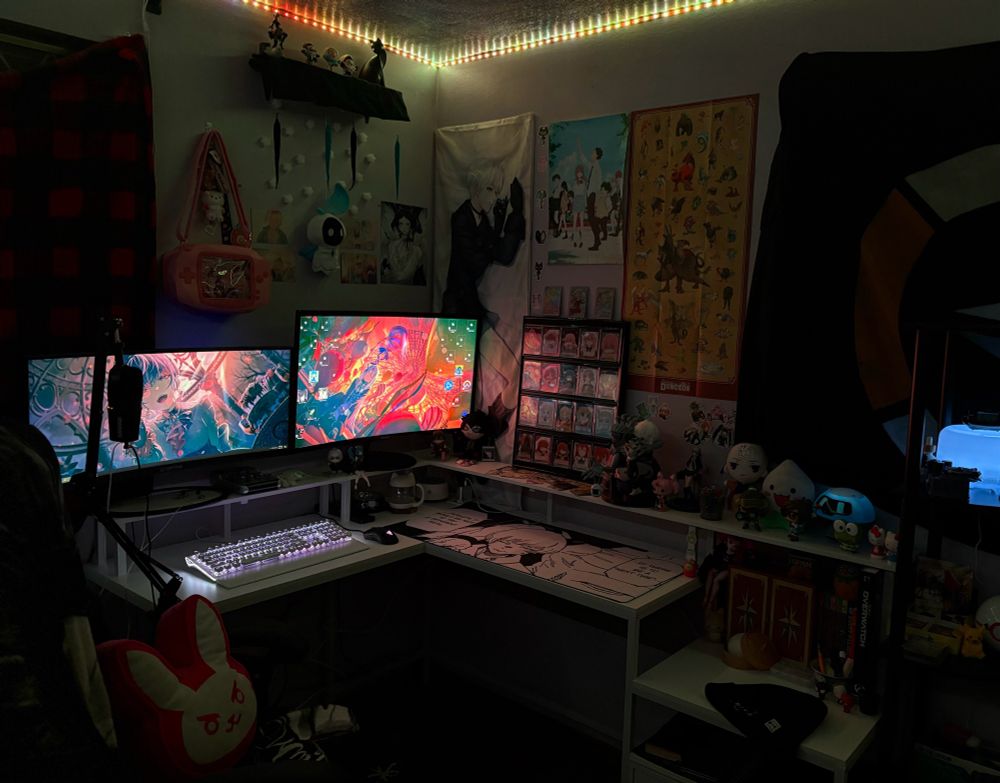 Picture of a desk. The desk has two monitors, and lots of different figures and collectibles displayed on it from several medias like anime, gaming, music, etc. the walls also have posters and stickers hung up. 