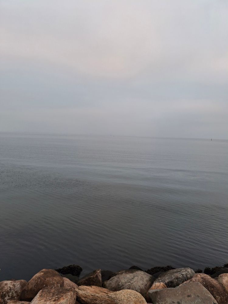 View over the open water. The sky and water are in different shafes of grey. Almost like they are one. The horizon is barely visible. At the bottom of the picture are a few big stones visible from the shore.