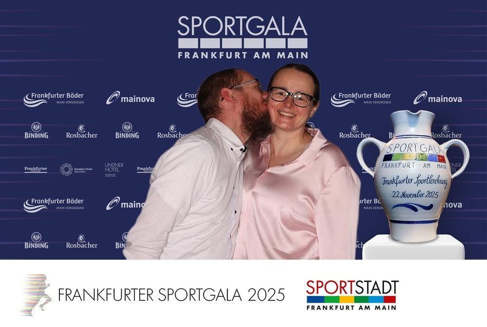 Mein Mann und ich in schicken Oberteilen (er weißes Hemd, ich rosane Bluse) vor einer blauen Wand, auf der in weisser Schrift Sportgala Frankfurt am Main steht. Sponsoren sind auch aufgeführt. Rechts unten ist ein für die Sportgala dekorierter, grauer Bembel reingephotoshopt. Ich schaue in die Kamera. Mein Mann küsst mir auf die Wange.