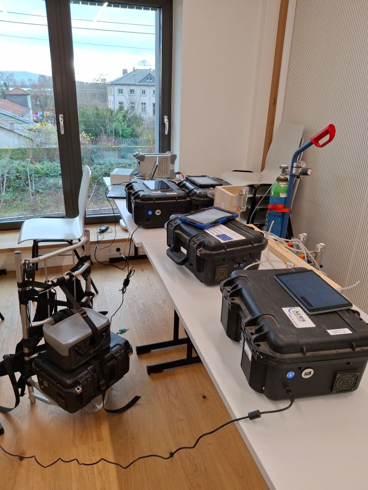 A row of tables in a lecture room. On the tables are 4 black suitcases (= analyzer) with tablets. One suitcase stands on the floor in a backpack like construction. In the right background, a gas cylinder with regulator can be seen. 
