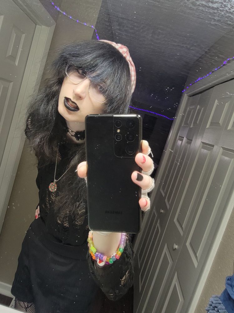 Janna in a very goth all black ripped outfit holding phone to mirror with black and blue dyed hair and black lipstick