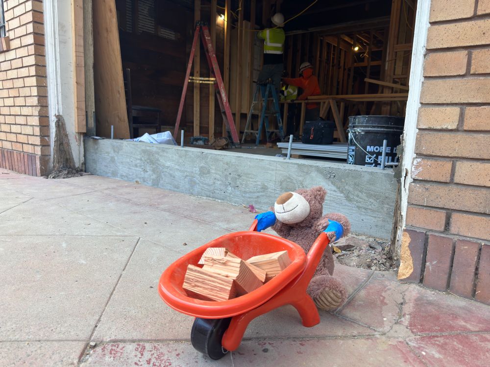 Toy bear with a toy wheelbarrow in front of a garage under construction.