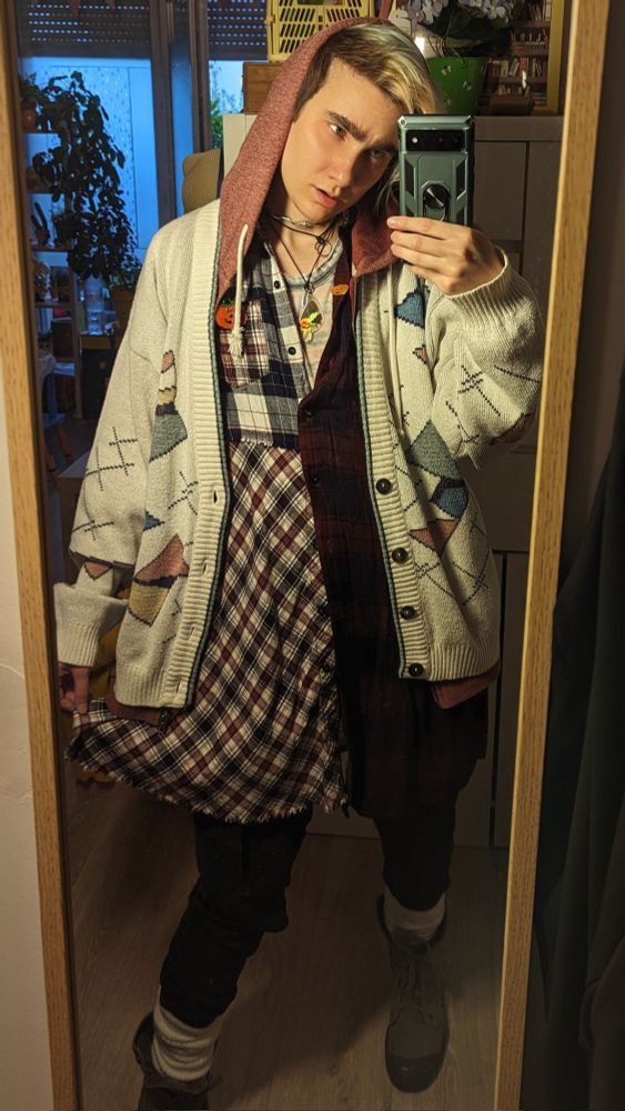 Selfie miroir dans mon appart. Je tire légèrement sur ma robe chemise à carreaux asymétriques blanc et bordeau d'un côté et bordeau et bleu foncé de l'autre, et je regarde mon portable avec un visage énervé. Je porte sous ma robe dont le col est ouvert un t'shirt rayé rouge et gris très délavé, par dessus j'ai un sweat zippé à capuche bordeaux, la capuche est sur ma tête. Par dessus je porte un large cardigan blanc cassé avec des motifs jacquard pastel éparses qui ressemblent à des rapiecement, un pantalon cargo noir, des guêtres blanc cassé, des bottines en cuir noir, 2 pins en forme de citrouilles oranges, un pendentif avec une aile de papillon brun, vert et orange et un choker fil de barbelé.