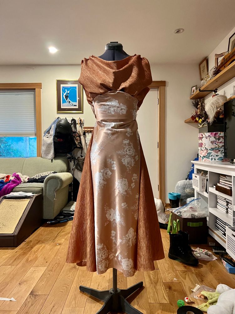 A dress is displayed on a dress form. It is two tone, with the main body of the dress being a shiny floral fabric in a warm toned gold. The accents are also a warm gold, but darker in tone. It has a draped cowl neckline, and side gores for additional volume in the skirt. 