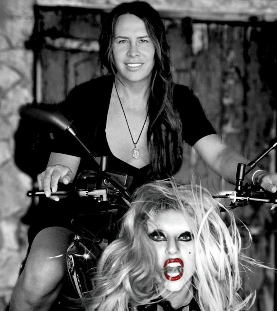 foto em preto e branco da Karla Sofia gascon sorrindo em cima de uma moto e na frente da moto tem a cabeça da lady gaga igual na capa do born this way 