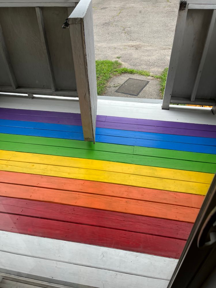 Our porch deck, painted with Pride flag colors, each color two planks wide, with the remainder painted white. Red is closest to the viewer (inside our kitchen), while purple is furthest away. The porch door is open, swinging inward. 