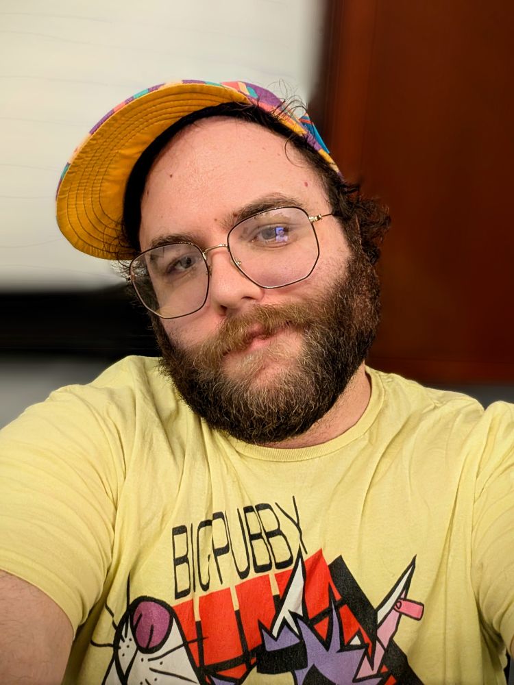 A white person with a beard, a purple hat, and a yellow shirt looking at the camera.