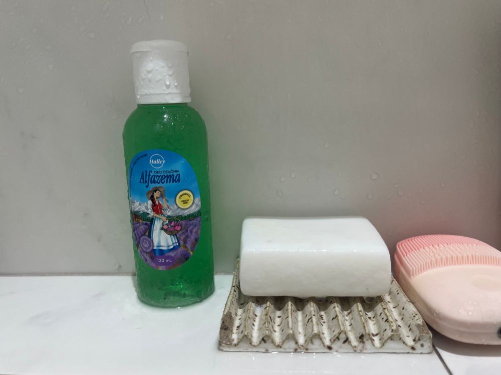 Imagem de dentro de um box de banheiro com um potinho de 120ml de deocolônia de alfazema cheio, ao lado direito uma saboneteira de cerâmica com um sabão de côco e ao lado uma esponja facial cor de rosa