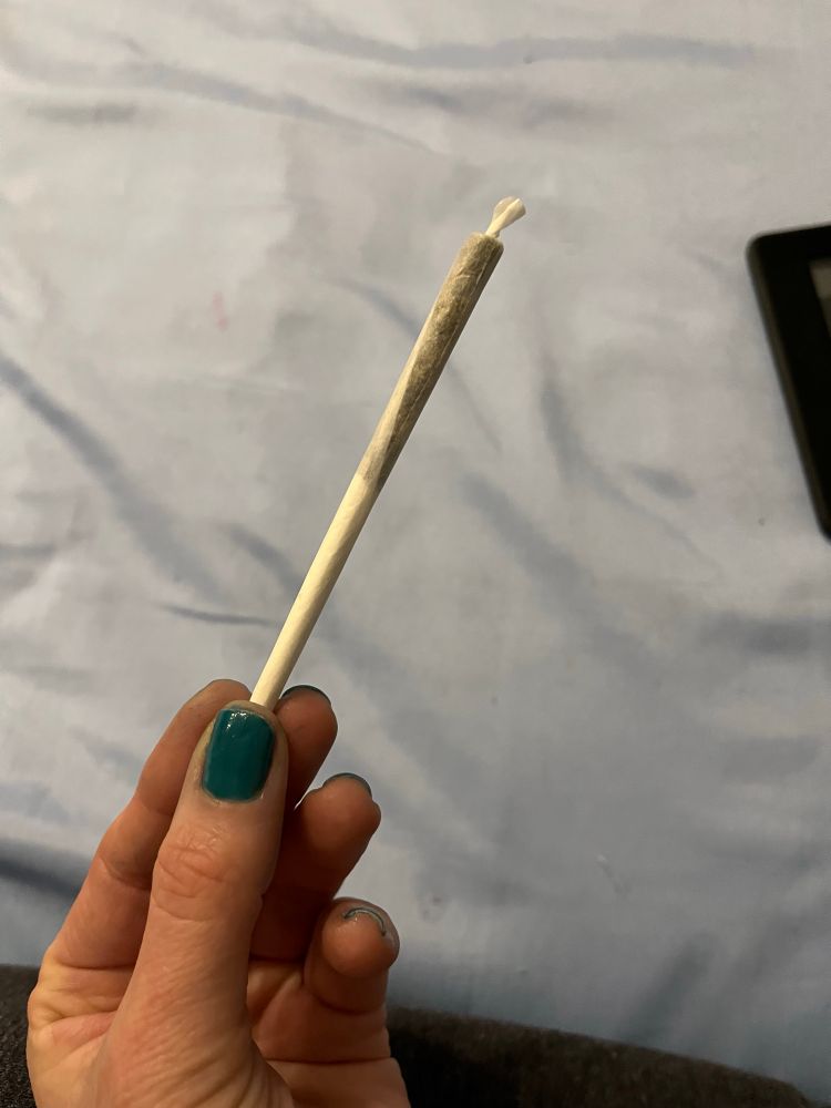 Minha mão esquerda, tenho pele branca a unha está curta e com esmalte verde escuro segurando um baseado fino com piteira de papel bem longa e a pontinha torcida, o fundo é um tecido azul claro