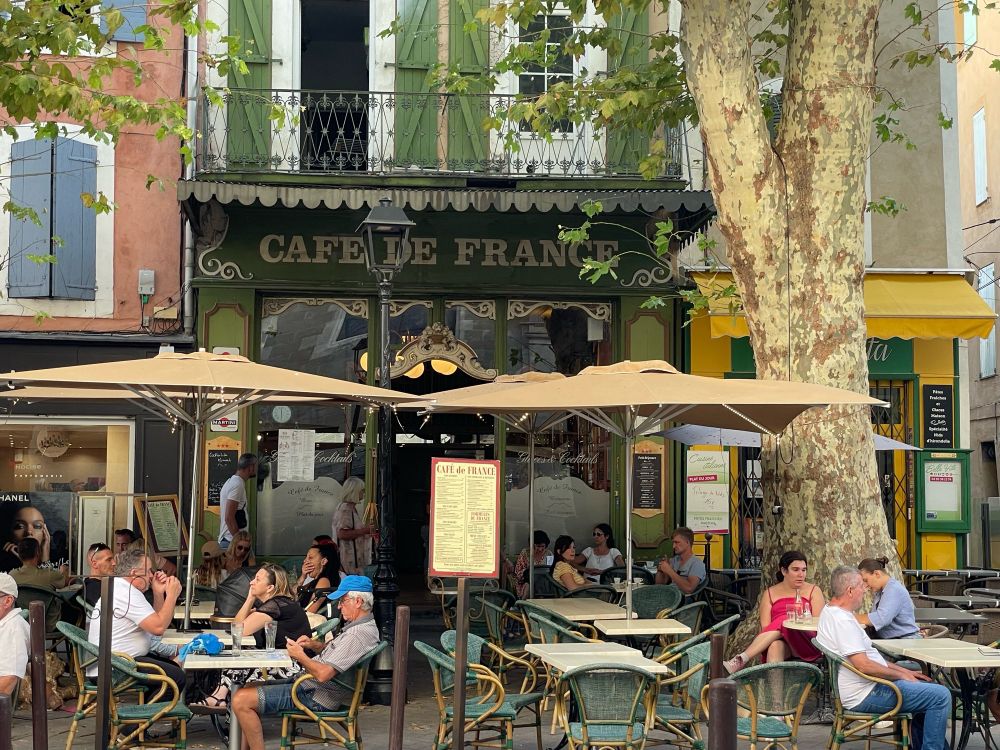 Zu sehen ist ein typisches altes französisches Café mit Sitzplätzen die unter großen Platanen sind