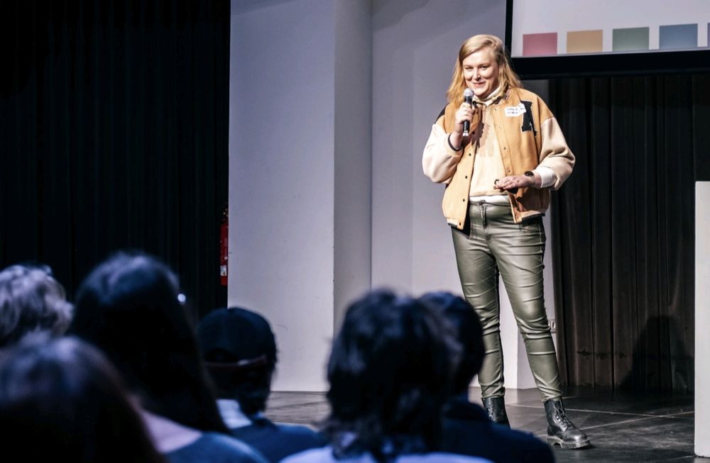 A blonde woman on a stage holding a microphone. She is talking to an audience in the shadows