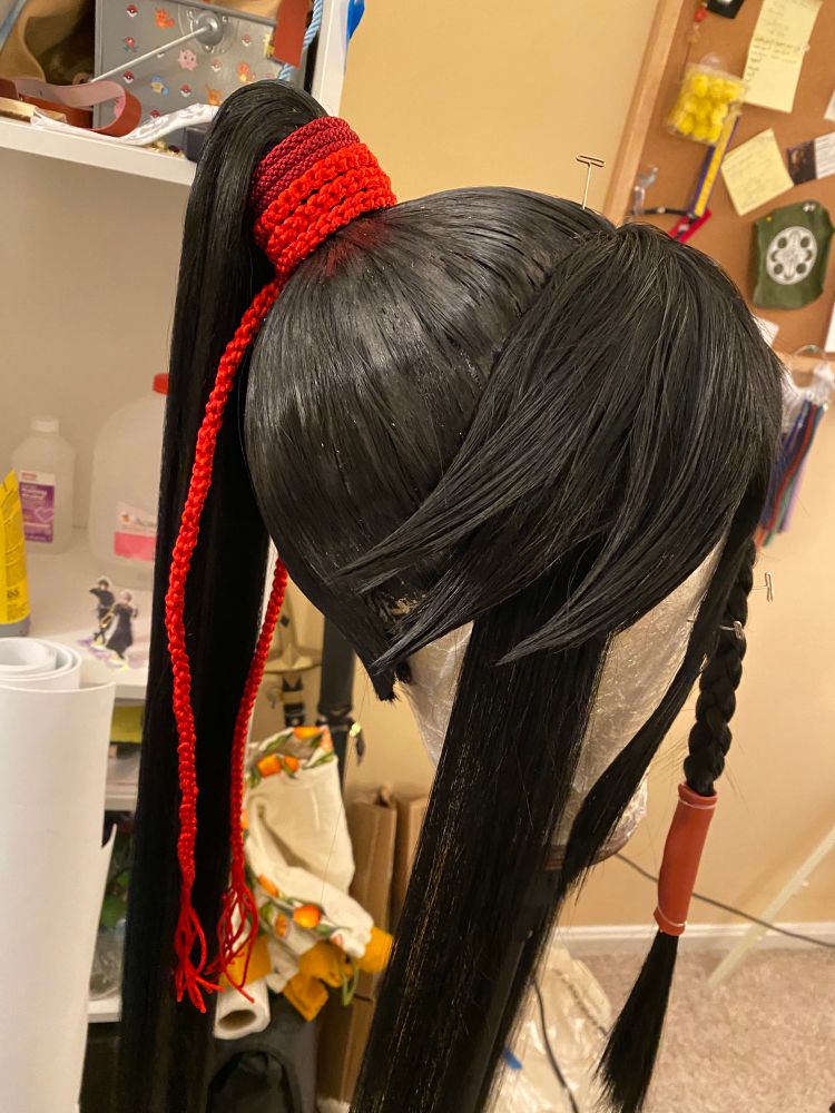 A black wig on a wig head as seen from the left profile. The wig is styled in a high ponytail with spikey bangs. The ponytail has two braids wrapped around it as hair ties. One is bright red and thicker; the other is darker red and thinner. Both are wrapped around the ponytail three times and the thicker one then dangles down on either side. Wig for Izuminokami Kanesada from Touken Ranbu.