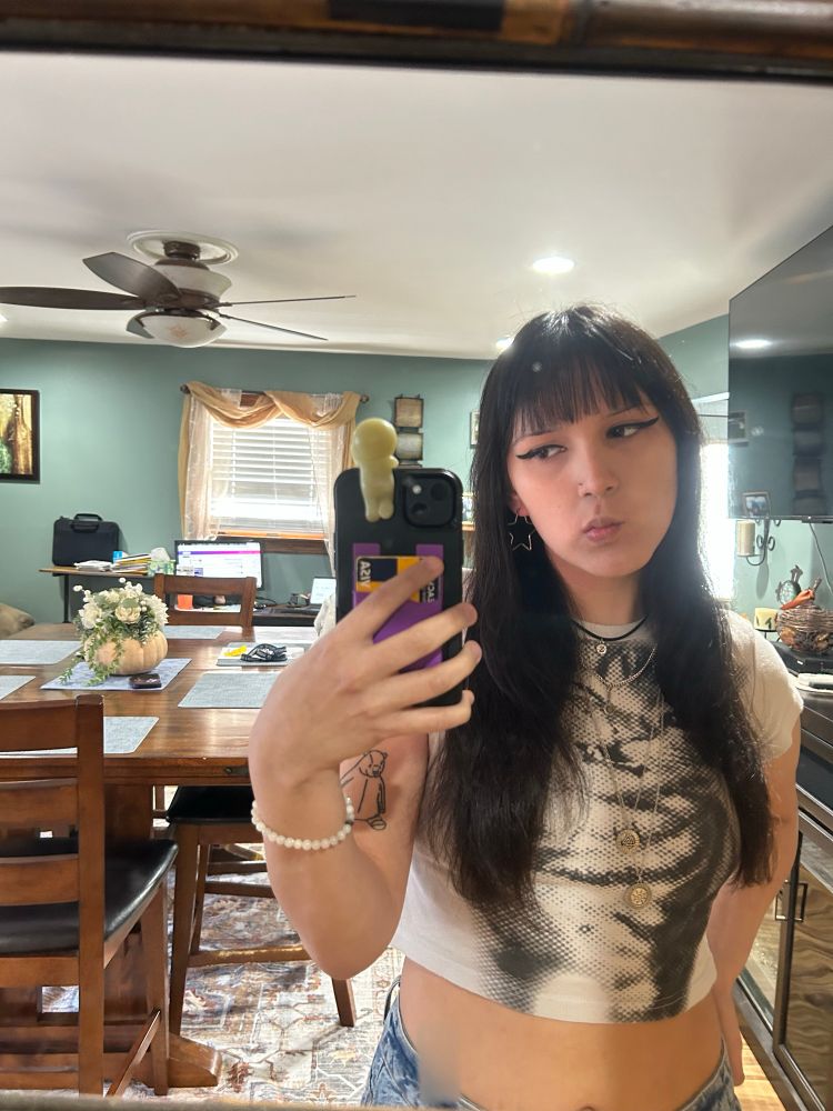 trans girl (me) with bangs and a djungelskog tattoo. shes wearing a silly cat shirt, star earrings, layered necklaces, and is thinking about her wife