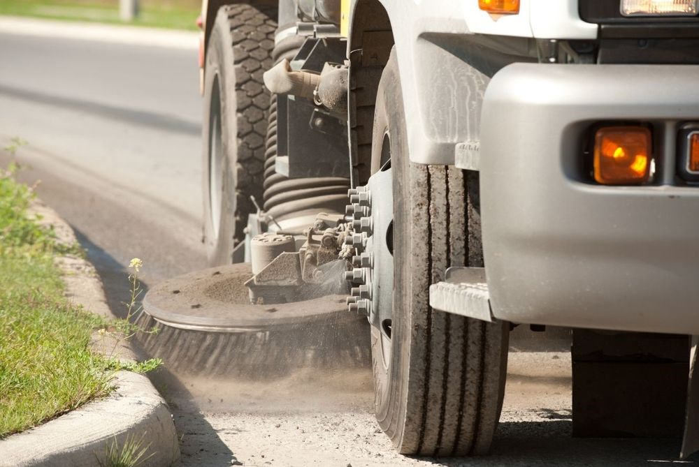 A close-up of a street sweeping vehicle next to a sidewalk. The broom is in motion and sweeping up debris. 