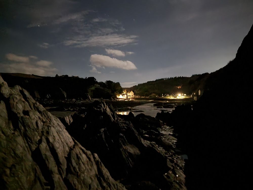 Night scene. Rocky beach in foreground, trees and well lit buildings behind. Quite clear night sky with a few clouds.