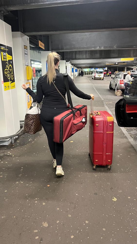 Iona stands at airport drop off with a red nord piano round her shoulder and red suitcase at hand with hair in a pony 