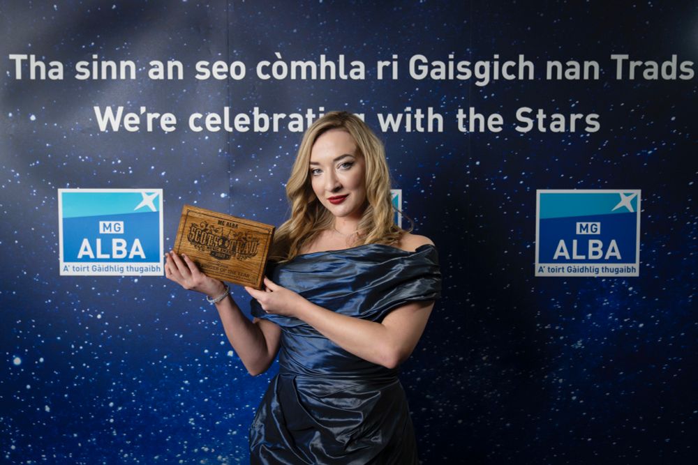 Iona Fyfe stands with a blue background with MG ALBA logo. Iona is wearing a blue dress and holding an award for the Scots Trad Music Awards