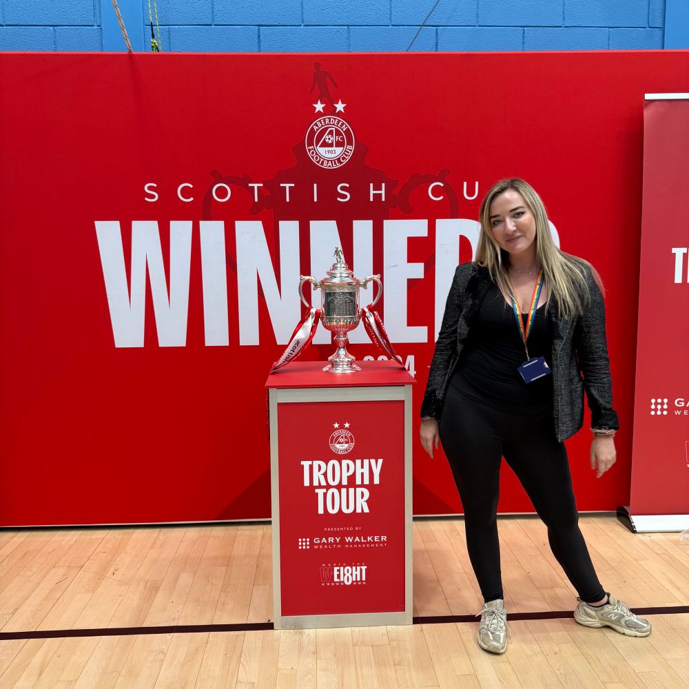 Iona stands next to the Scottish Cup Trophy with a background of red Aberdeen Football Club branding and a plinth that says Trophy Tour