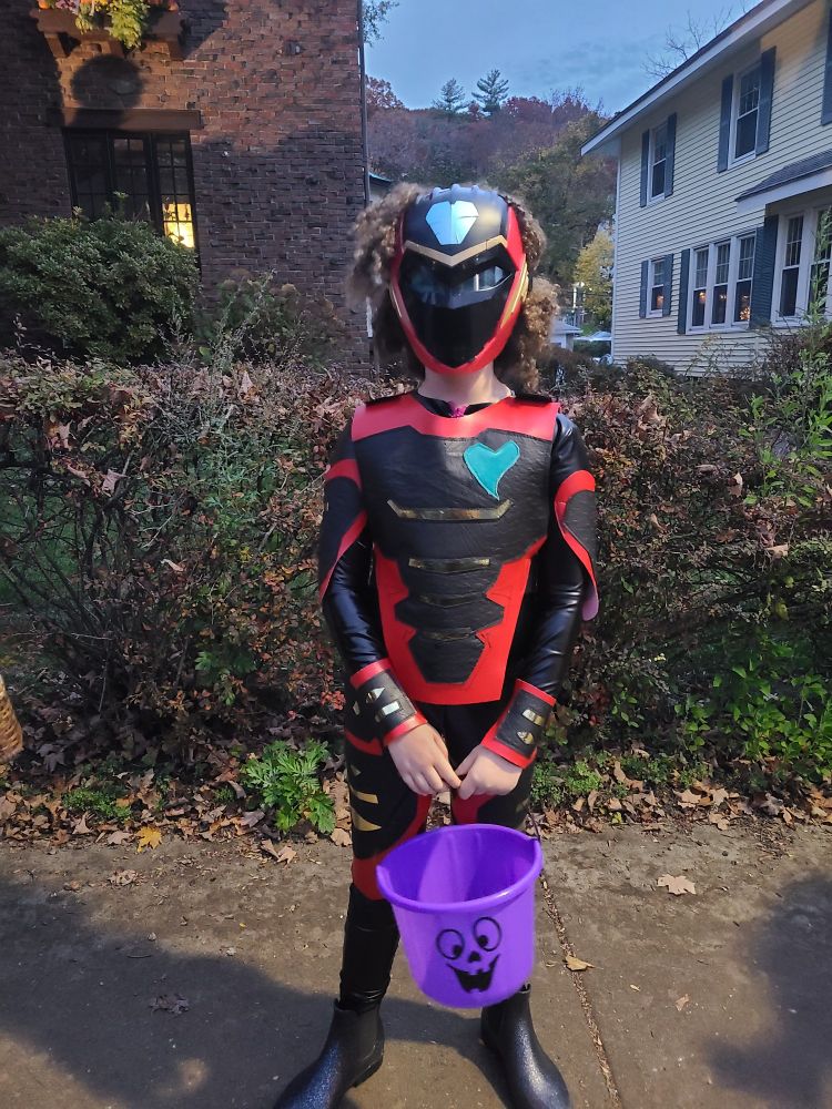 A child in a custom made ironheart costume stands in front of a bush with a purple ghost pail. 
