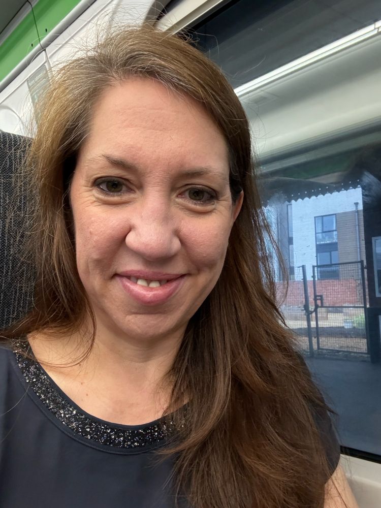 An image of a white woman with long brown hair. She is smiling at the camera whilst siting on a train. 