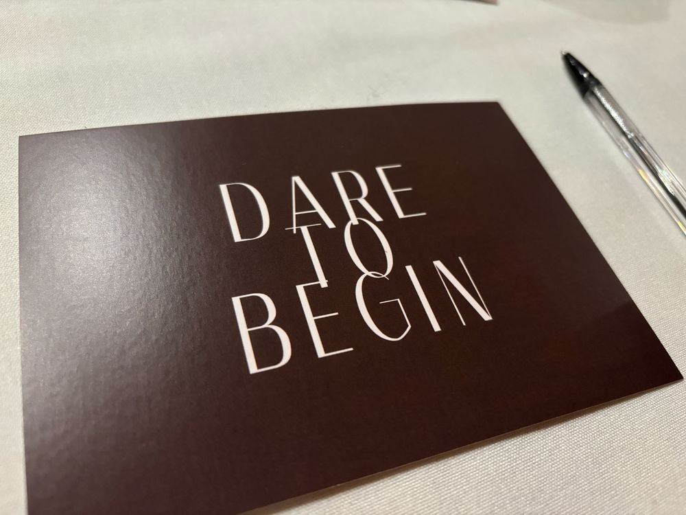 An image of a postcard on a table. The postcard says DARE TO BEGIN