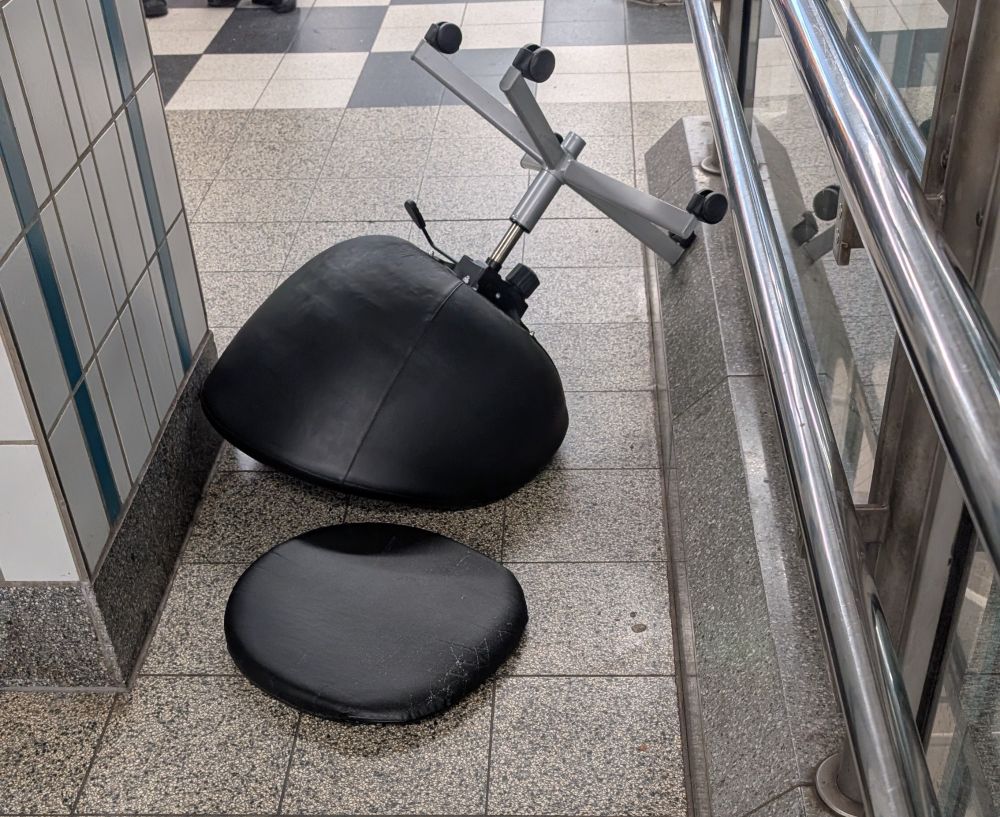 Schwarzer Stuhl mit Rollen unten liegt Kopfüber auf Fliesenboden zwischen gefliester Säule und Glasgeländer, die fünf schwarzen Rollen stehen schräg ab und zeigen nach unten 